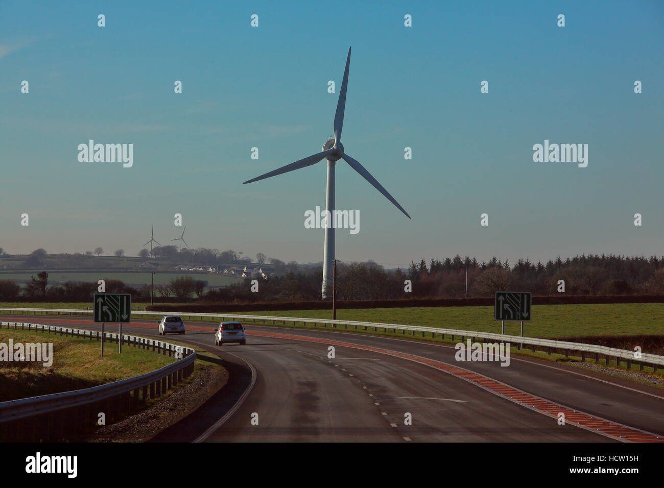 Eine schöne zweispurige mit Beschilderung und eine riesige Windkraft Turbine im Hintergrund vor einem strahlend blauen Himmel. Stockfoto