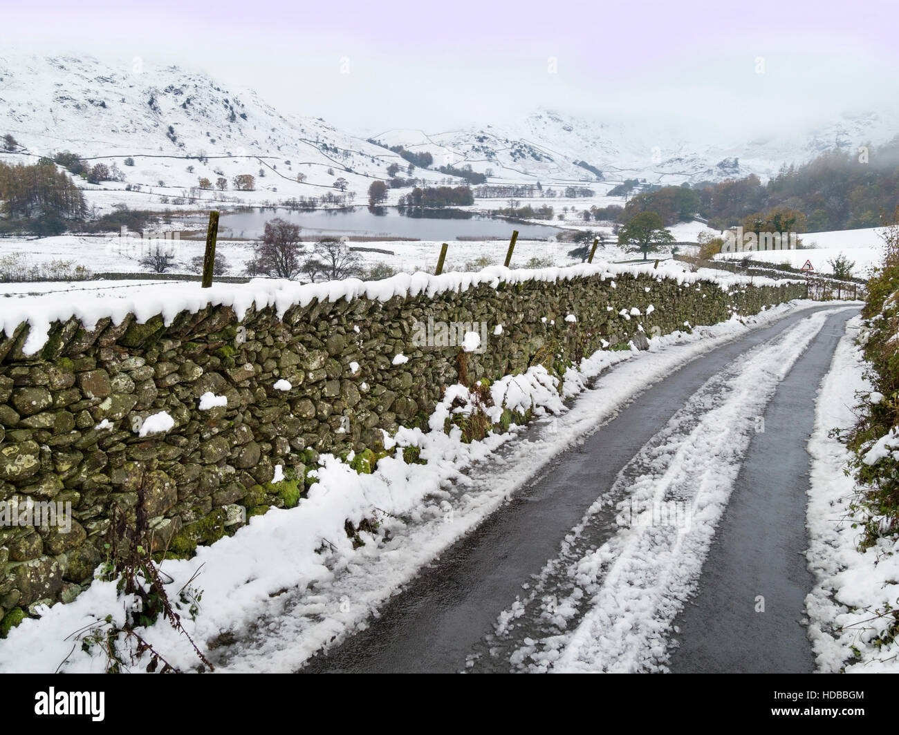 Verschneite Straße mit trockener Steinmauer in Little Langdale mit tarn Beyond, English Lake District, Cumbria, England, Großbritannien. Stockfoto
