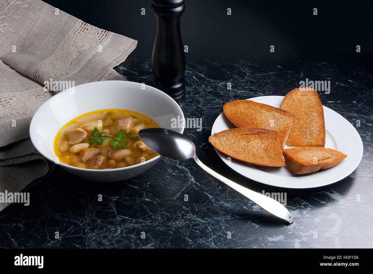 Bohnensuppe in weiße Schüssel mit Metalllöffel auf einem schwarzen Stein Hintergrund. Einige toast auf weißen Teller und schwarze Mühle für Pfeffer auf braunem Tuch. Stockfoto