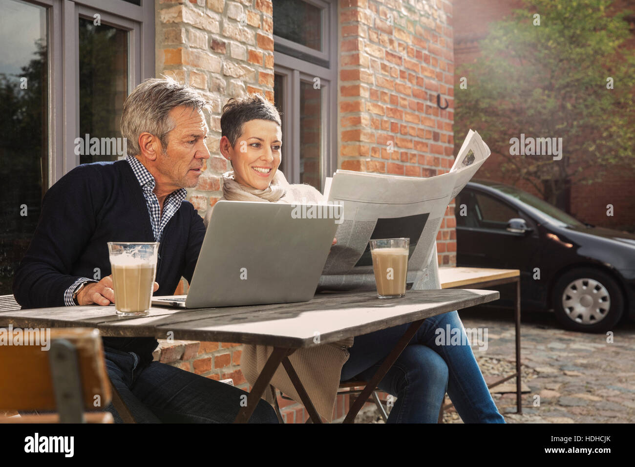 Reifer Mann und Frau liest Zeitung sitzend mit Laptop im Hinterhof Stockfoto