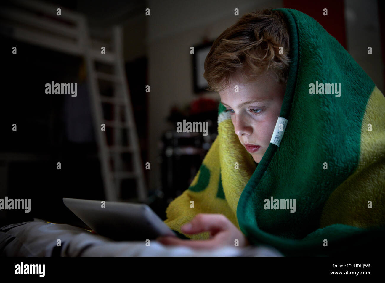 Ein Junge mit einem Handtuch über dem Kopf auf seinem Bett liegend mit einem digitalen Tablet. Stockfoto