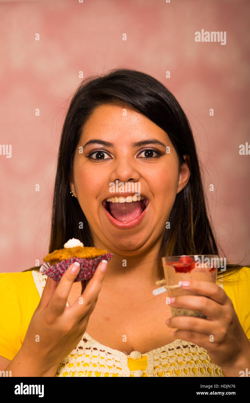 Brünette Frau mit köstlichen braunen farbigen Muffin mit Sahne-topping, Glas Mousse in der anderen Hand, Lächeln und bereit, ein Biss, Gebäck-Konzept Stockfoto