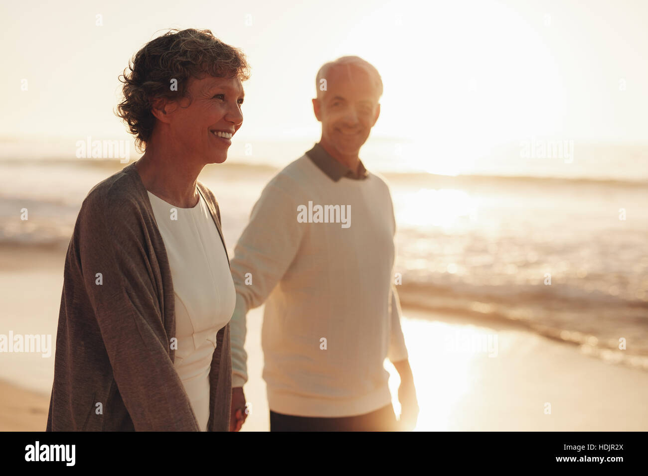 Lächelnde senior Frau mit ihrem Ehemann zu Fuß am Strand bei Sonnenuntergang. Älteres Paar zu Fuß entlang der Küste. Stockfoto