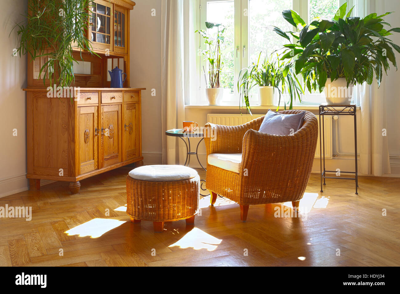 Sonniges Wohnzimmer in warmen Farben mit gemütlichen Wicker Sessel und Hocker, Vintage Schrank, Pflanzen und großen Fenstern, nostalgische Stockfoto