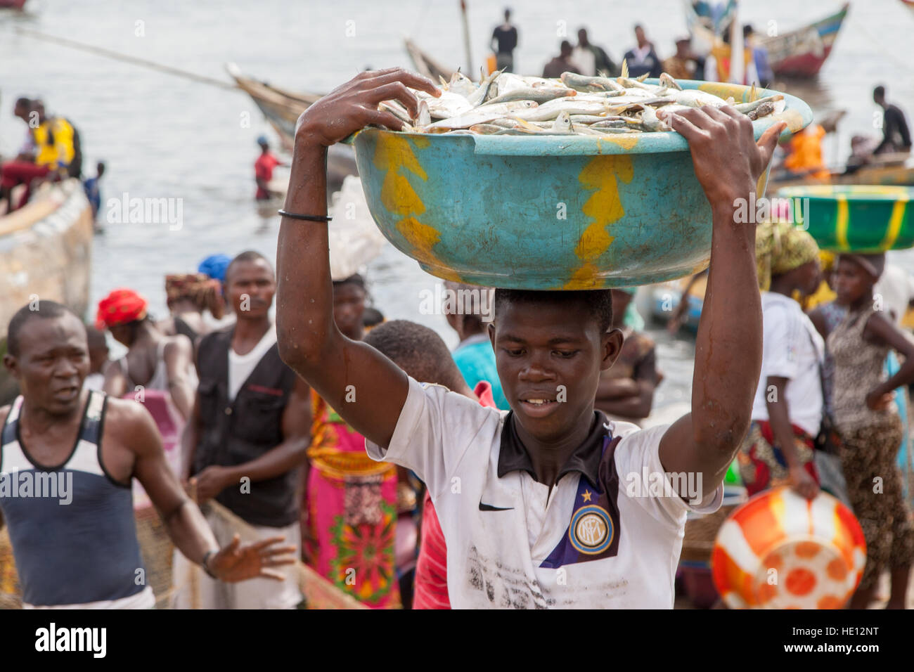 Junge Männer, die schwere Schalen mit Fischen von Boot zu Ufer tragen. Frauen warten darauf, dass weitere Fischerboote ankommen. Tombo, Sierra Leone Stockfoto