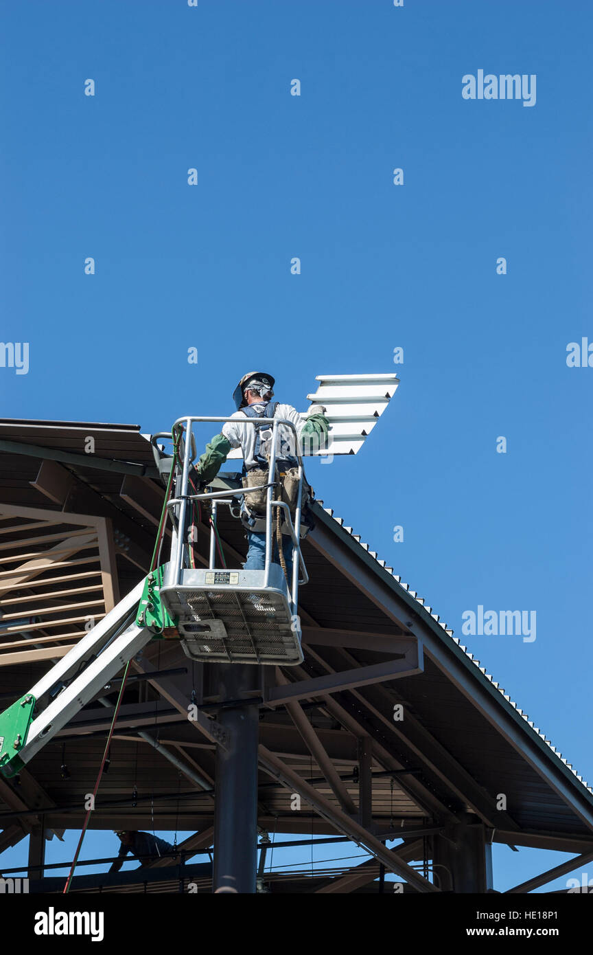 Ein männliche Dach Auftragnehmer in einem Boom Lift Korb tragen persönlicher Schutzausrüstung installiert Metall Dachplatten in einem Industriegebäude Stockfoto