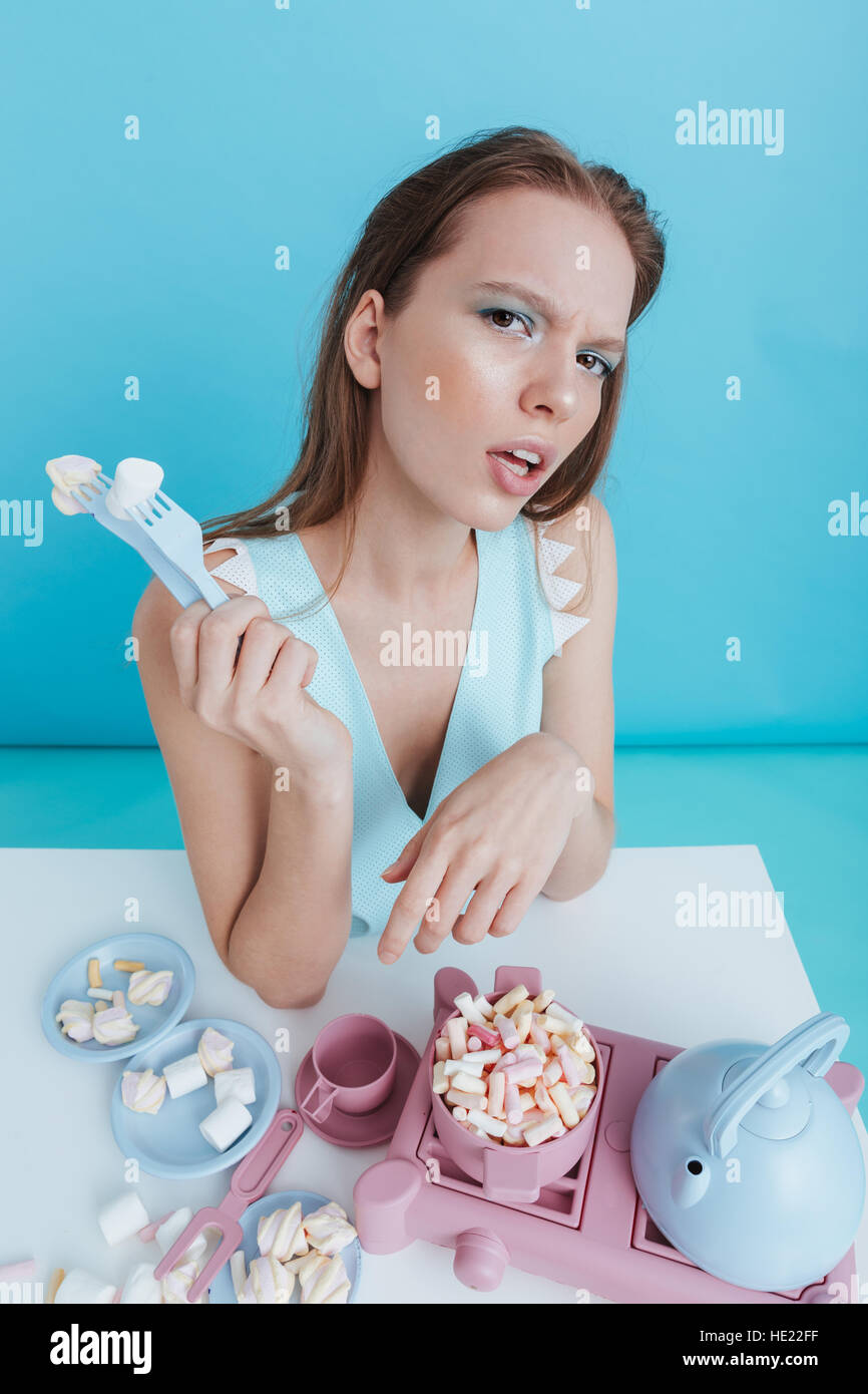 Unglücklich verwirrt junge Frau, die Verwendung von Plastik Gabeln zum spielen und Essen Marshmallows auf blauem Hintergrund Stockfoto