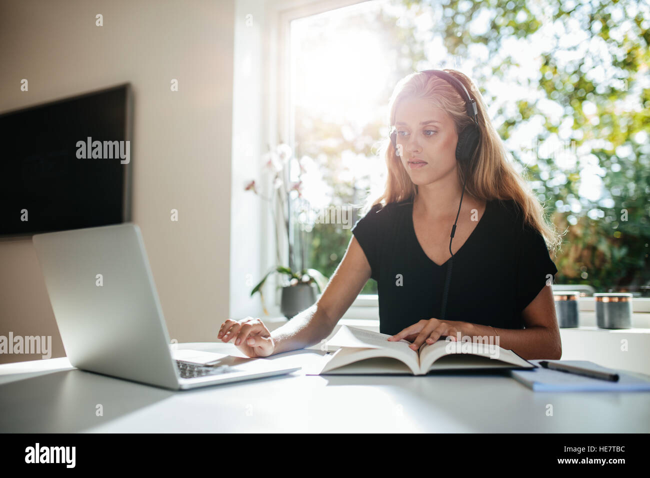 Junge Studentin Studium zu Hause. Junge Frau mit Buch mit Laptop am Tisch sitzen. Stockfoto