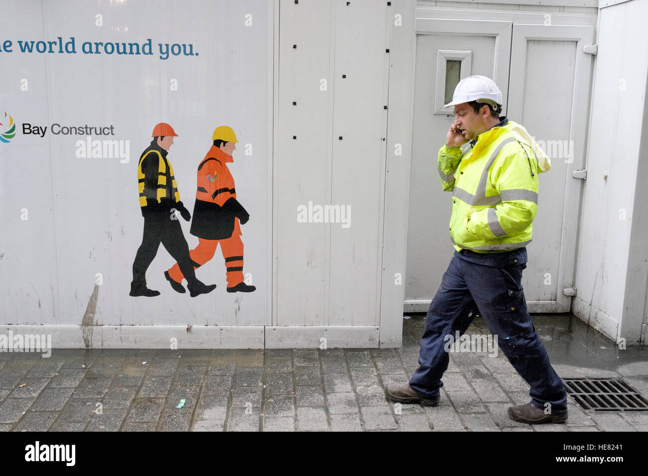 Ein Arbeiter chats auf seinem Handy, während er hinter einer temporären Wand zeigt Bilder der Arbeiter zu Fuß in die andere Richtung geht Stockfoto