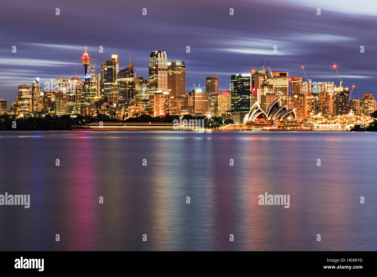 Sydney CBD Citylight und Wasser durch den Hafen bei Sonnenuntergang. Helle Ausleuchtung der Wolkenkratzer und australischen Wahrzeichen unter unscharfen bewölkten Himmel. Stockfoto