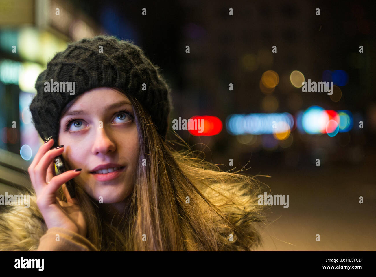 Glückliche Mädchen in Hut und Mantel telefonieren mit Handy lächelnd gegen Nachtlichter der Stadt im winter Stockfoto