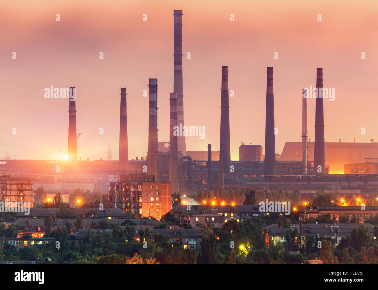 Gebäude der Stadt auf dem Hintergrund der Stahlfabrik mit Schornsteinen bei Sonnenuntergang. Metallurgische Fabrik mit Kamin. Schwerindustrie Stockfoto