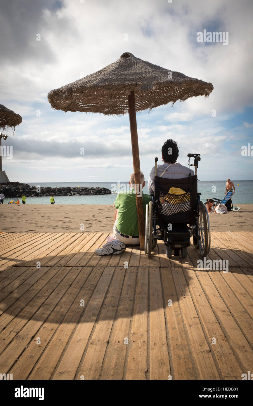 Mann und Woaman an einem Strand, Blick auf das Meer unter einem Sonnenschirm zu sitzen, ist Frau im Rollstuhl Stockfoto