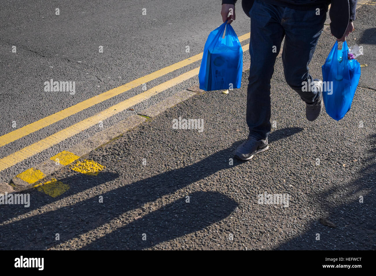 Mann trägt beleuchtet Einkaufen in blauen Plastiktüten, wieder einen Schatten zu schaffen. Angrenzend ist eine Straße Bordstein und doppelte gelbe Linien Stockfoto
