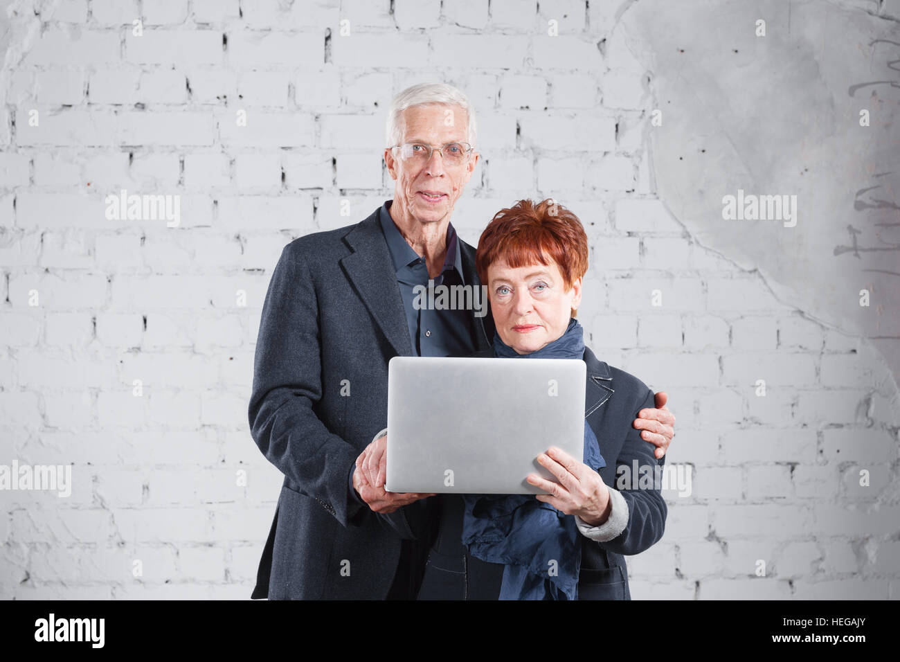 Alte Leute halten einen Laptop und über das Internet zu kommunizieren. Glücklich lächelnd Opa Oma paar stehende kuscheln zusammen auf weiße Ziegel Hintergrund isoliert. Stockfoto