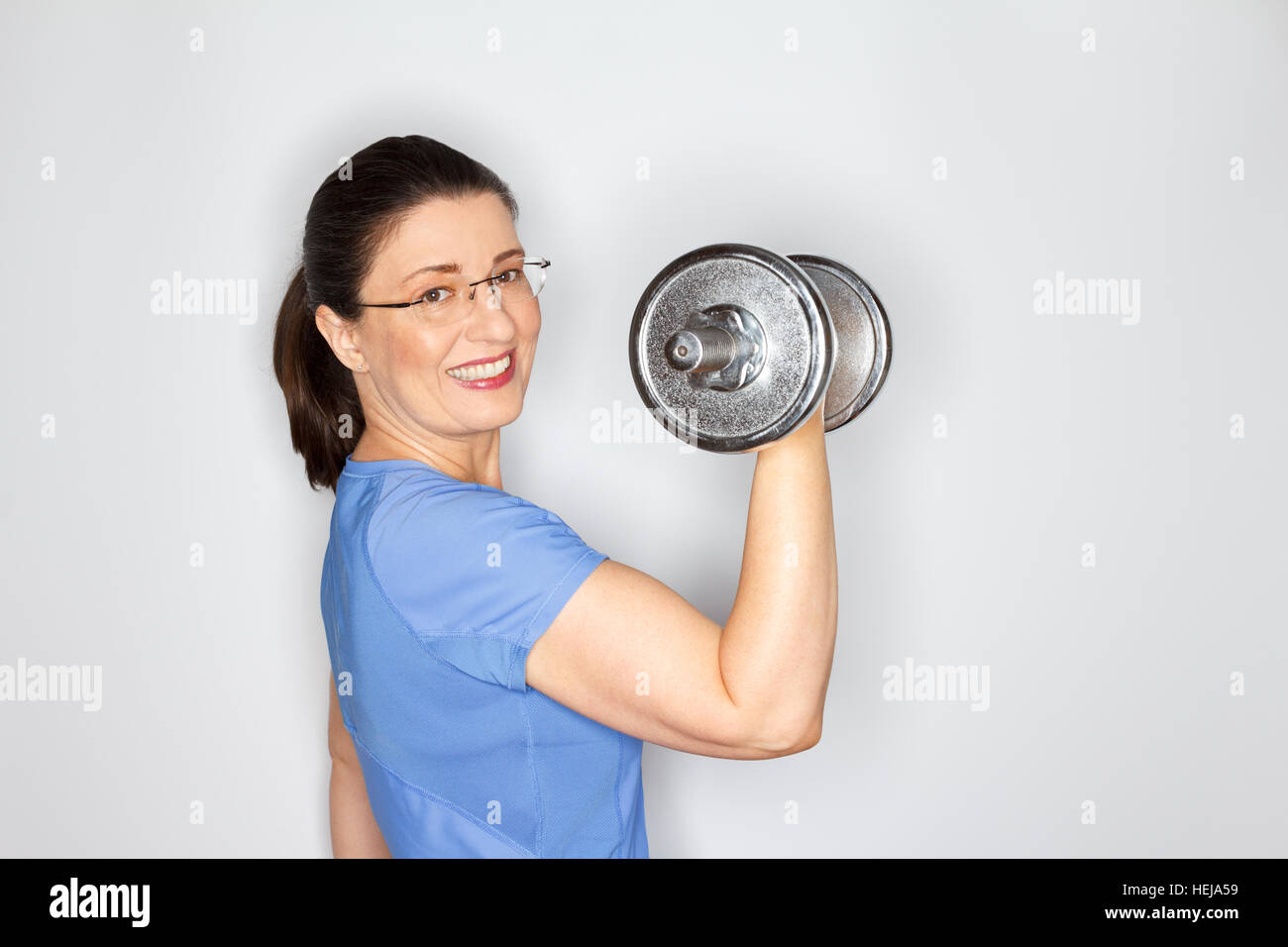 Glücklich und stolz mittlere gealterte Frau mit Brille und im blauen T-shirt heben Gewichte, hellgrauen Hintergrund, Textfreiraum, Exemplar Stockfoto