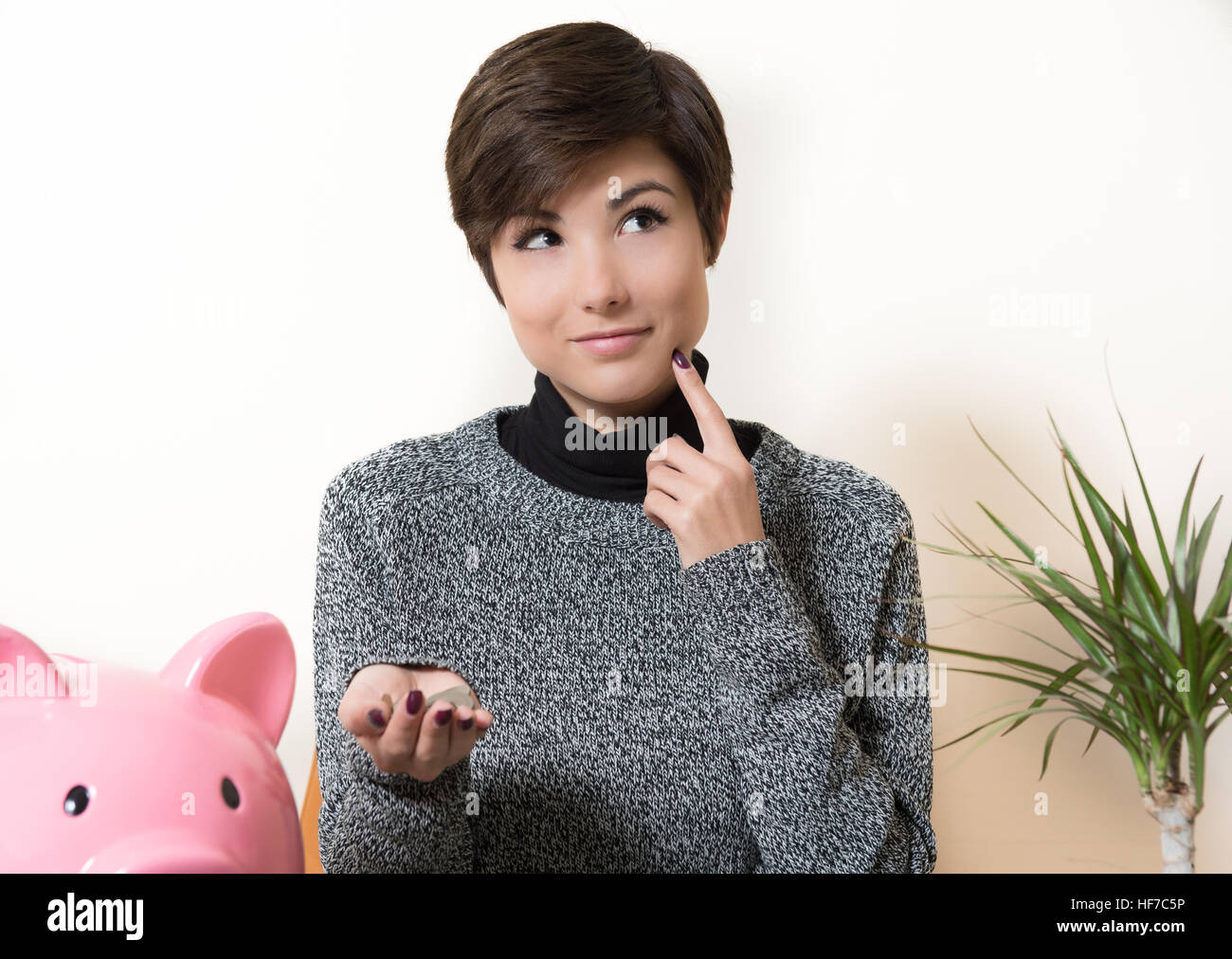 Junge Frau lächelt und zum Nachdenken über Einsparungen, Münzen in die Hand und rosa Sparschwein auf desktop Stockfoto