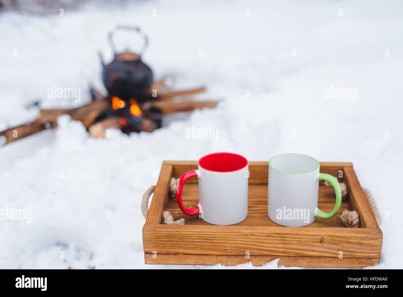 Romantische Winter-Picknick. Zwei Tassen auf einem Holztablett im Schnee. Kupferkessel über dem offenen Feuer auf Hintergrund unscharf. Wasserkocher auf Brennholz. Kopieren Sie sp Stockfoto