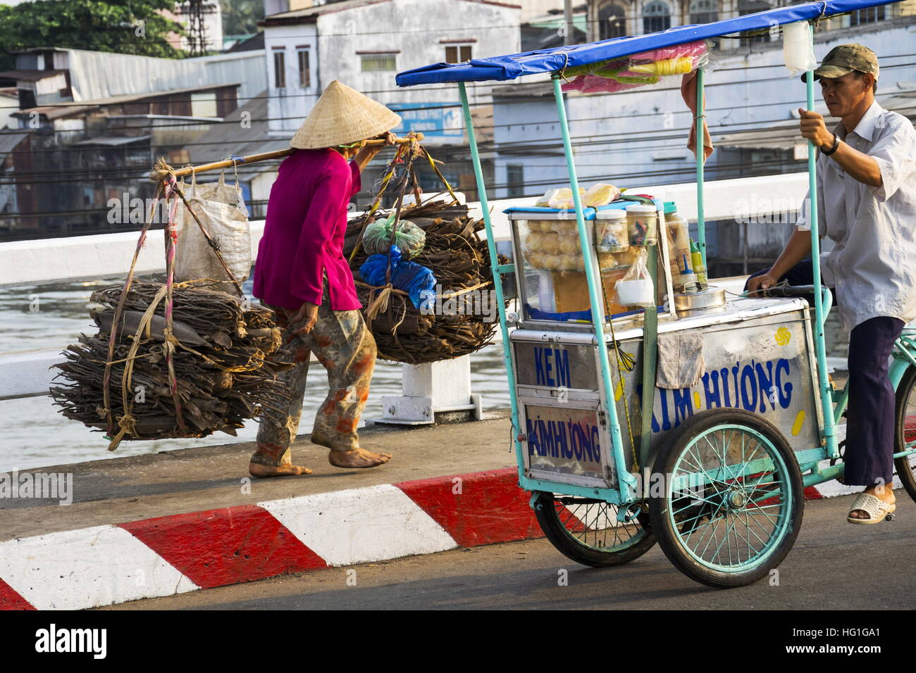 Barfuß Vietnamesisch Reife Frau in konischen asiatischen Hut tragen Holz an viel befahrenen Straße in der Nähe von chinesischen Viertels in My Tho, Vietnam. Stockfoto