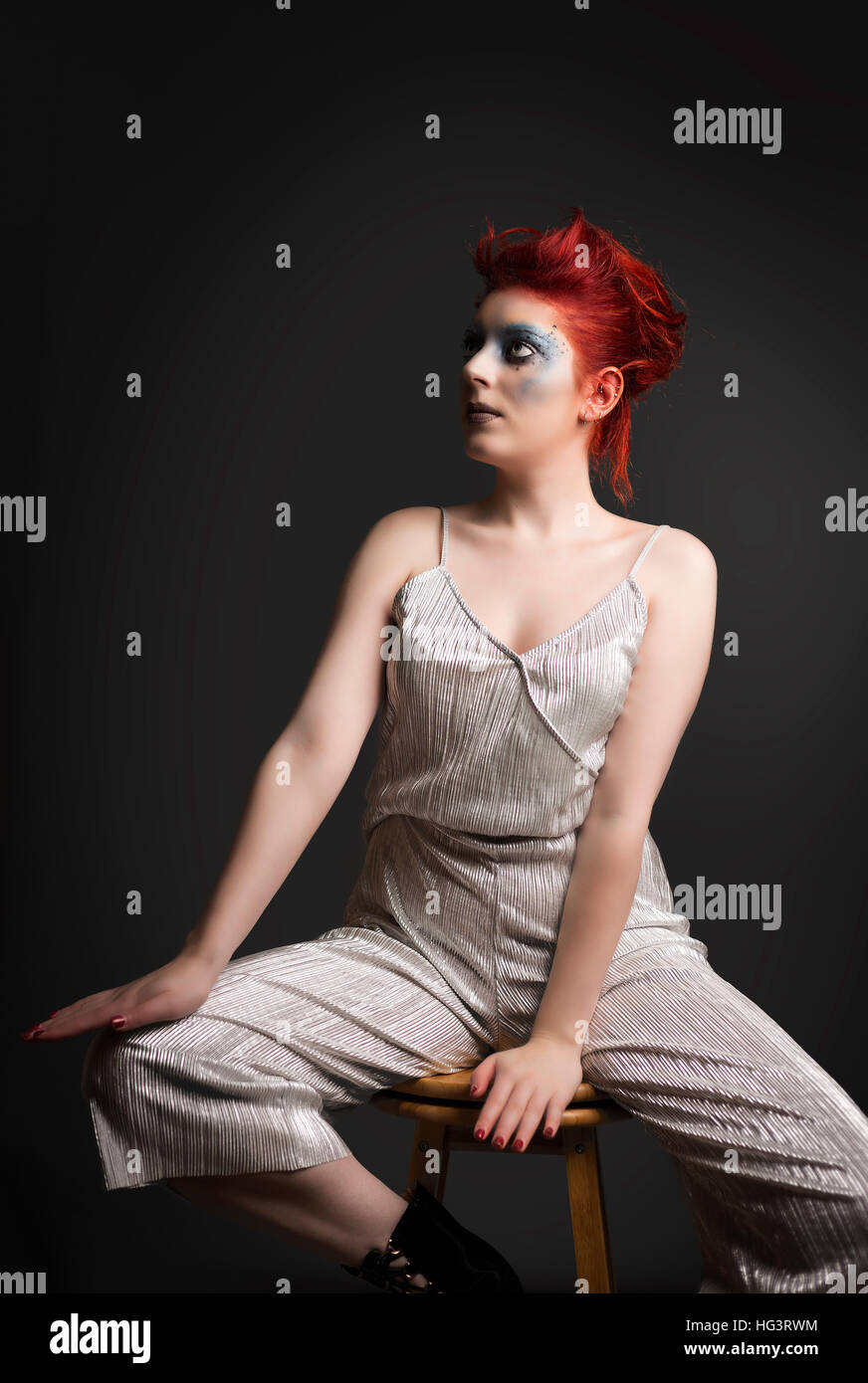 Roten Haaren Modell mit blauen kreative Make-up trägt Silber Overall, gesetzt auf hölzernen Schemel Stockfoto