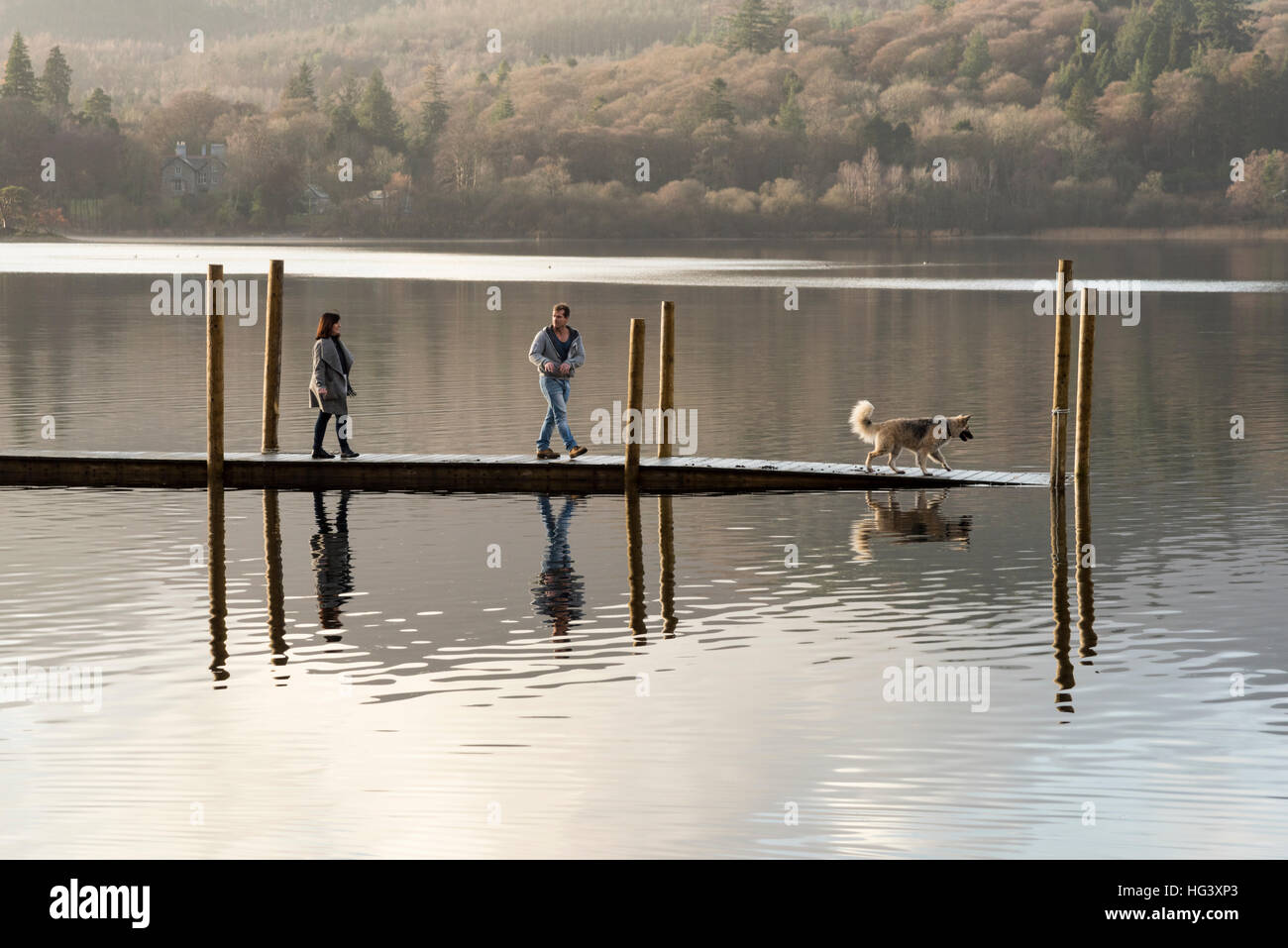 Zwei Personen mit einem Hund zu Fuß auf einem Holzsteg oder Liegeplatz auf Derwentwater Keswick Cumbria Uk im Wintersonne Stockfoto