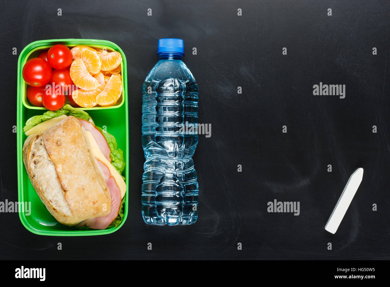 Sandwich, kleine Tomaten, Mandarine in Kunststoff Lunch-Box und eine Flasche Wasser an schwarze Tafel. Stockfoto