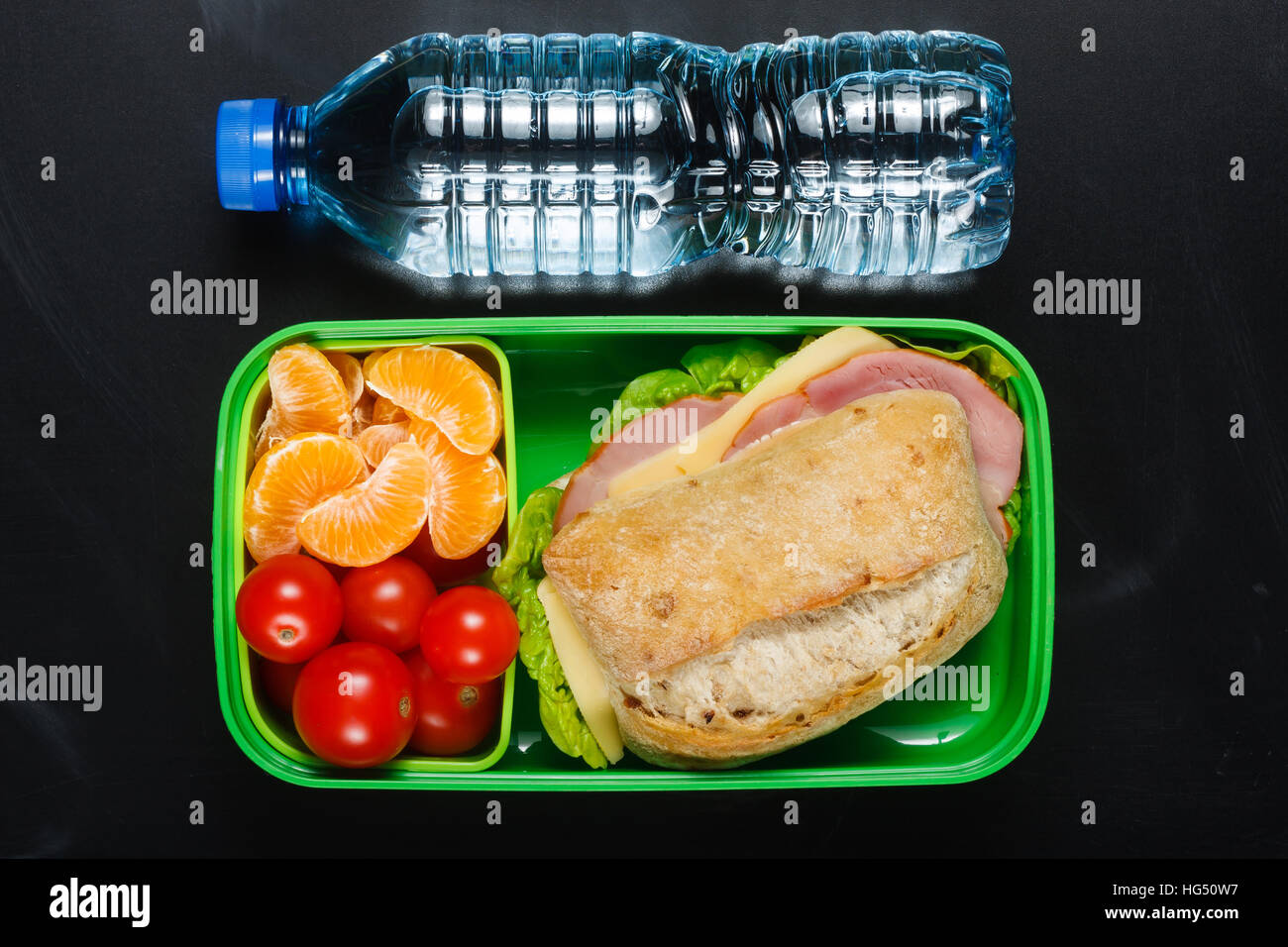 Sandwich, kleine Tomaten, Mandarinen in Kunststoff Lunch-Box und eine Flasche Wasser an schwarze Tafel. Stockfoto