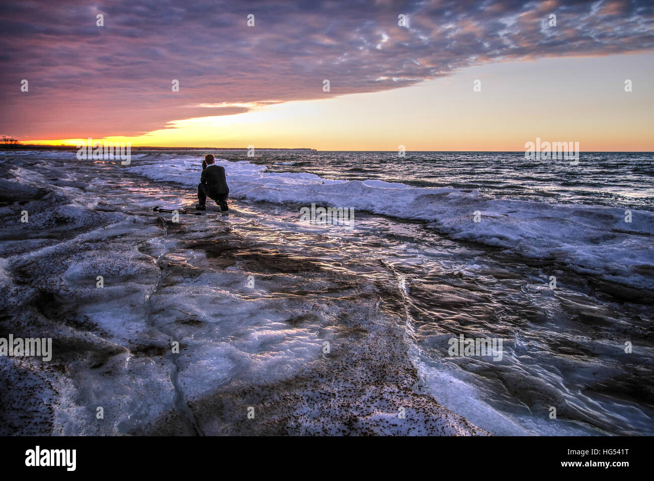 Männlichen Fotografen auf einem zugefrorenen See. Junge männliche Fotografen kniend auf der gefrorenen Küste des großen Sees bei Sonnenuntergang. Stockfoto