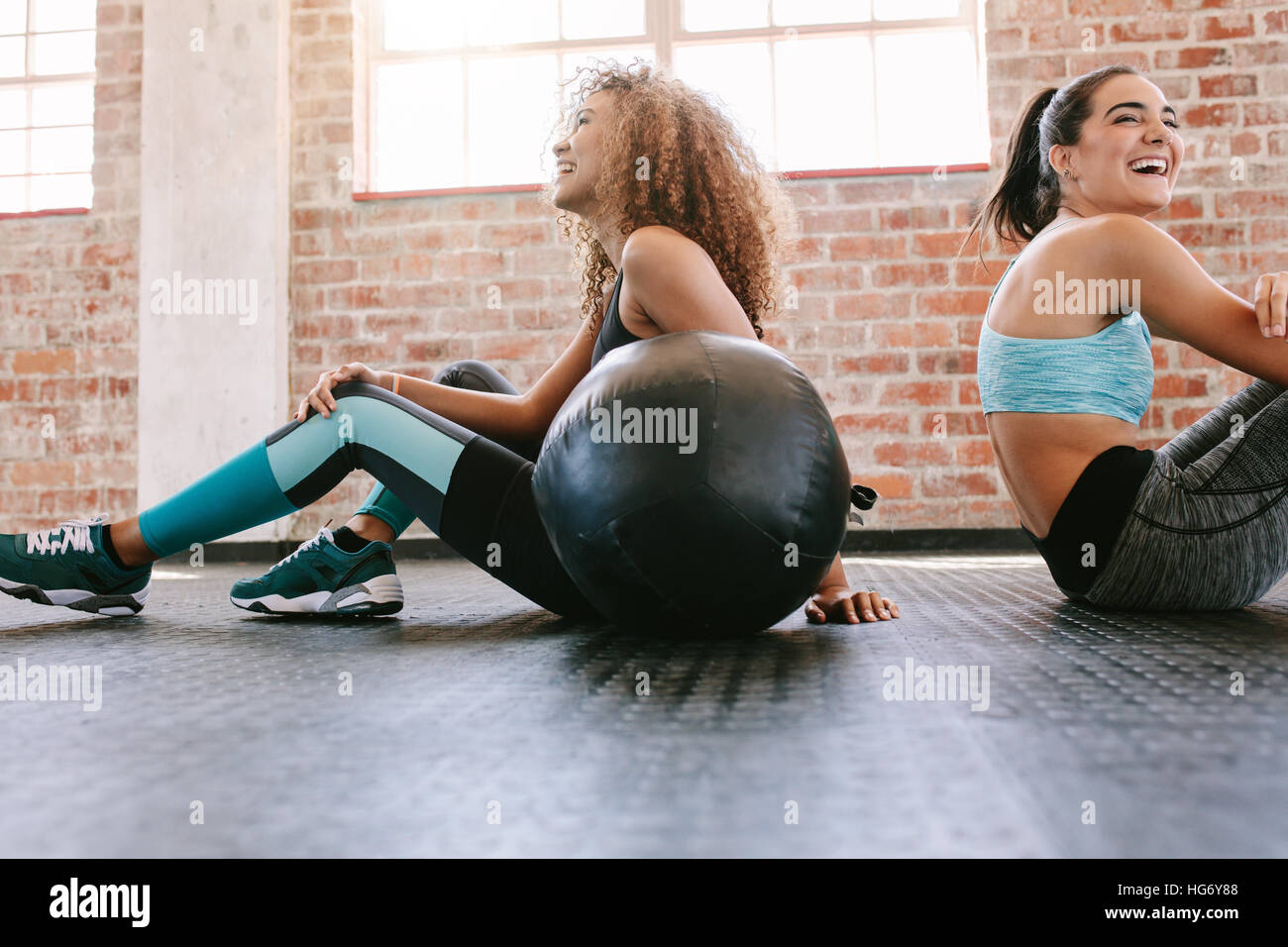 Glückliche junge Freundinnen sitzen im Fitnessstudio Stock mit Medizinball und lächelnd. Zwei junge Frauen, die eine Pause vom Training. Stockfoto