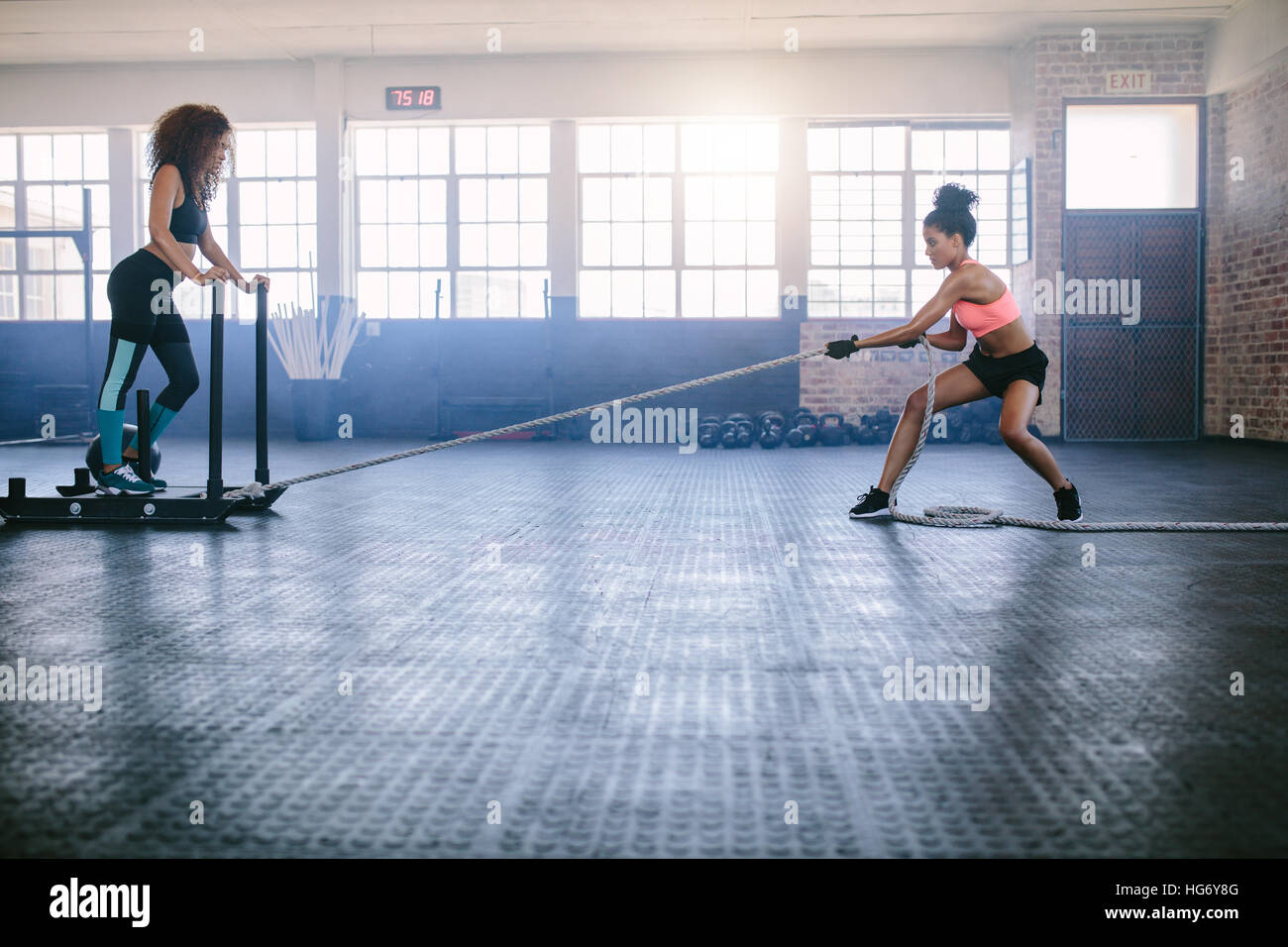 Fitness-Schlittenzug mit Frau auf Schlitten stehend. Zwei Frauen, die intensives körperliches Training im Fitnessstudio machen. Stockfoto