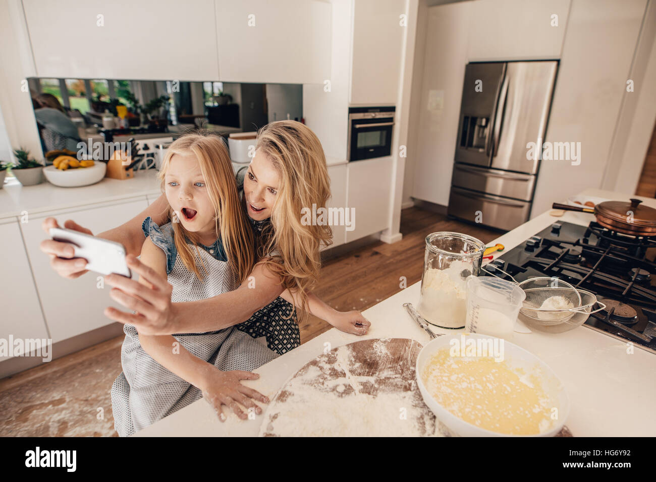 Glückliche junge Mutter nehmen Selfie mit ihrer Tochter in der Küche. Junge Frau und kleine Mädchen, die Zubereitung von Essen in Küche nehmen Selbstbildnis mit mobilen pho Stockfoto