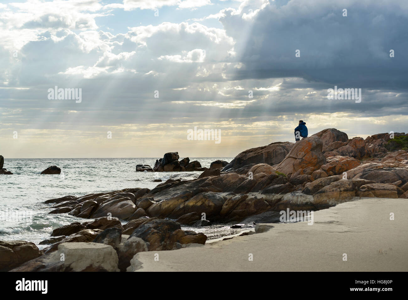 Ein Mann sitzt auf den Felsen an einem Strand, Blick auf den Ozean mit Göttern Strahlen Sonnenlicht durch Wolken Stockfoto