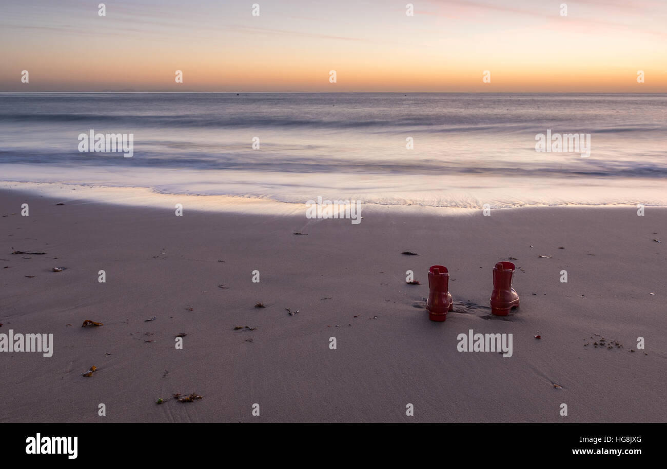 rote Gummistiefel am Ufer zurückgelassen bei Sonnenaufgang am Strand Stockfoto