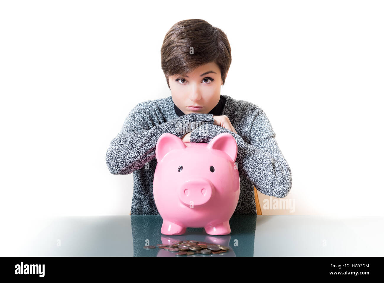 Junge Frau nachdenklich über Einsparungen, Münzen und rosa Sparschwein auf Desktop, isoliert weißen Hintergrund Stockfoto