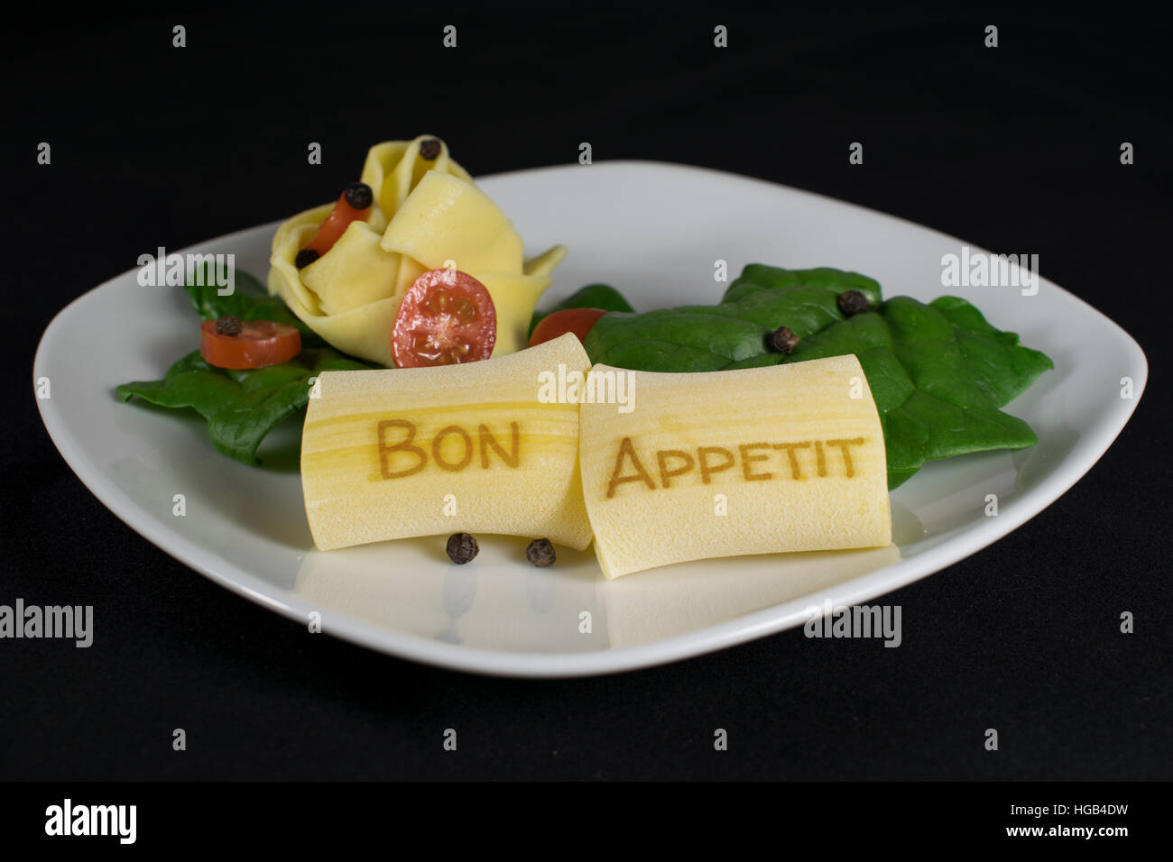 Auf einem weißen Teller sind 2 Nudeln mit dem Schriftzug "Bon Appetit" im Vordergrund. Im Hintergrund sind weitere Nudeln garniert mit Blattspinat, Stockfoto