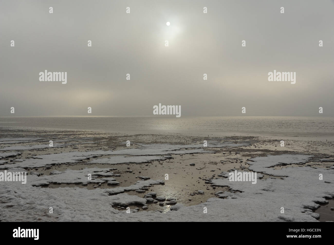 Kleine weiße Sonne durch graue Wolken über dem Sandstrand mit Meereis am Rande der cal Meerwasser, St Annes, Lancashire, UK Stockfoto