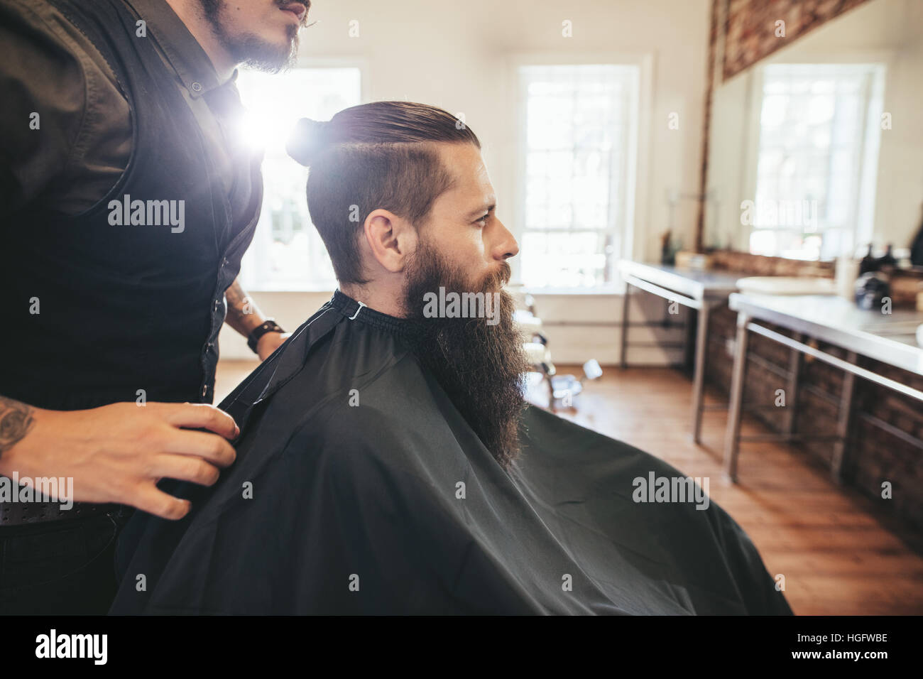 Seitenansicht Schuss von schöner bärtiger Mann in einem schwarzen schneiden Haare Kap im Barbershop, mit Friseur steht. Mann sitzt im Friseursalon. Stockfoto