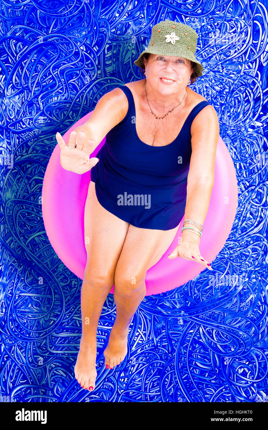 Ältere Dame in einem stilvollen Hut und Badeanzug schwimmend auf einem Rohr in das Schwimmbad entspannen Sie sich auf einen heißen Sommertag nachschlagen zu winken in die Kamera, Konz Stockfoto