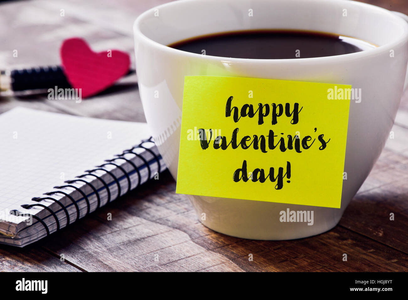 Nahaufnahme von eine gelbe Haftnotiz mit dem Text happy Valentines Day, dass Sie mit einer Tasse Kaffee auf einem rustikalen Holztisch, neben einem Notebook, eine erneute verbunden Stockfoto