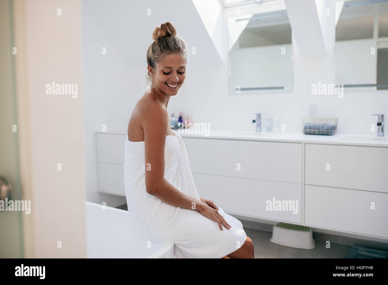 Lächelnde junge Frau sitzt auf Badewanne im Badezimmer nach Bad. Junge weibliche Modell in Handtuch gewickelt. Stockfoto