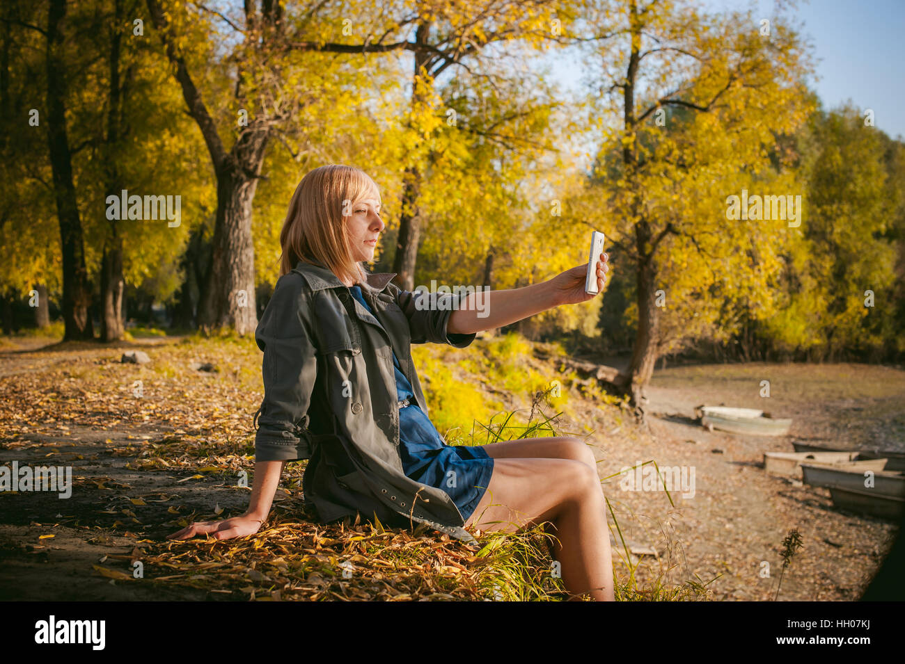 hübsche blonde Mädchen in Turnschuhe, Nylons und Mantel. fotografiert auf  einem Smartphone Selfie, sitzt am Ufer, auf gelbe Blätter, genießen Wärme  AUT Stockfotografie - Alamy