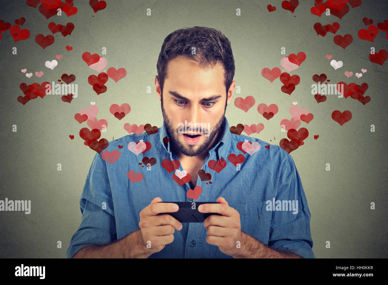 Porträt schöner junger schockiert Mann senden empfangen von Liebe Sms-Nachricht auf dem Handy mit roten Herzen wegfliegen isoliert auf graue Wand backg Stockfoto