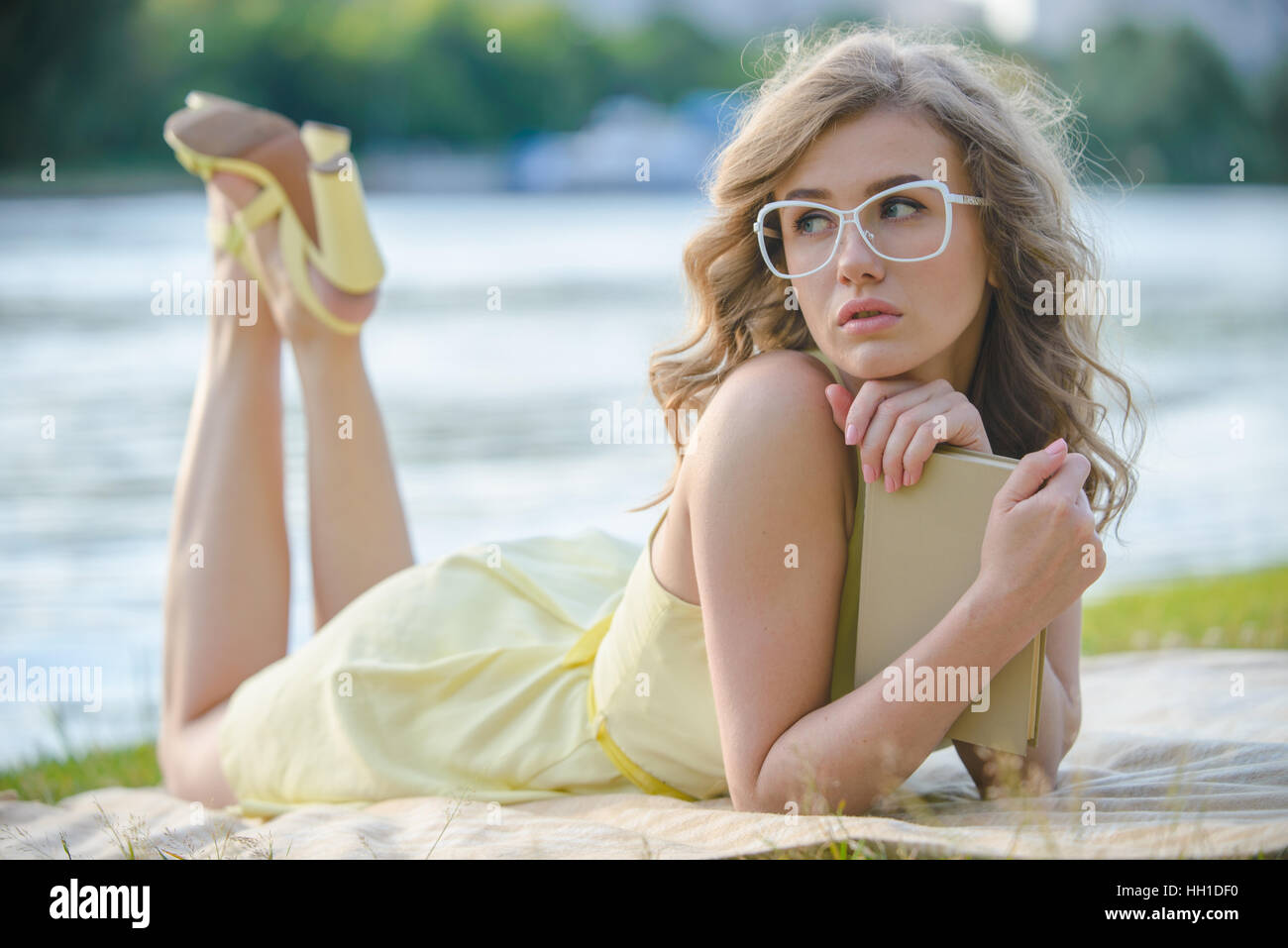 Junge Frau mit Buch am Ufer, gelbes Kleid, Brille, verträumt, modische Stockfoto