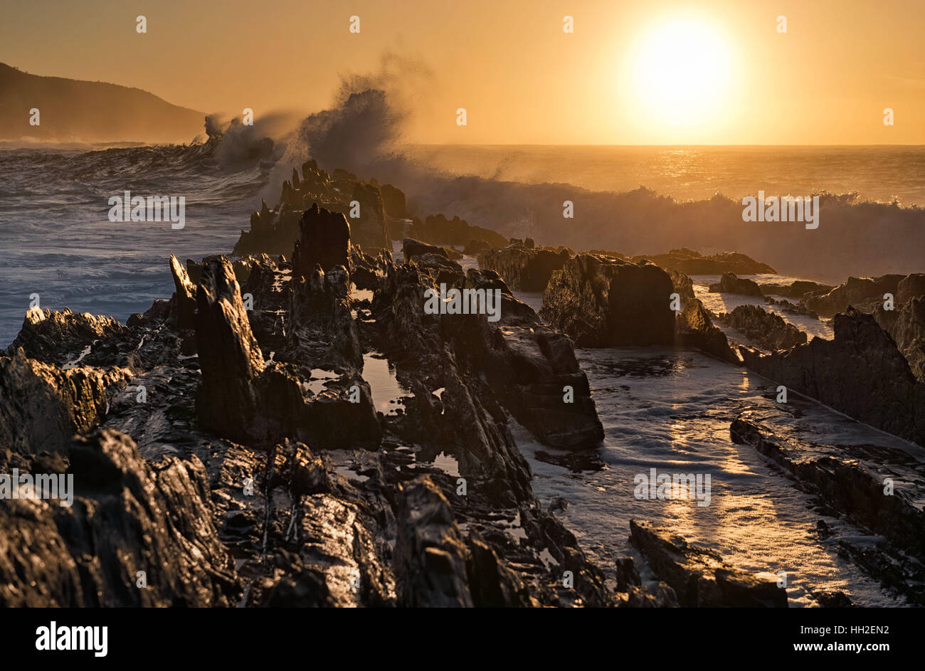 Wellen zum Absturz gegen schroffe Felsen während des Sonnenuntergangs am Storms River, Südafrika Stockfoto