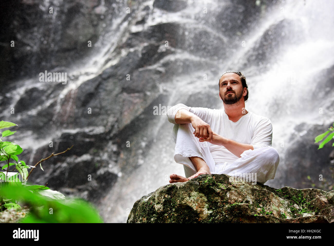 Mann sitzt in Meditation Yoga auf Felsen am Wasserfall im Regenwald Stockfoto