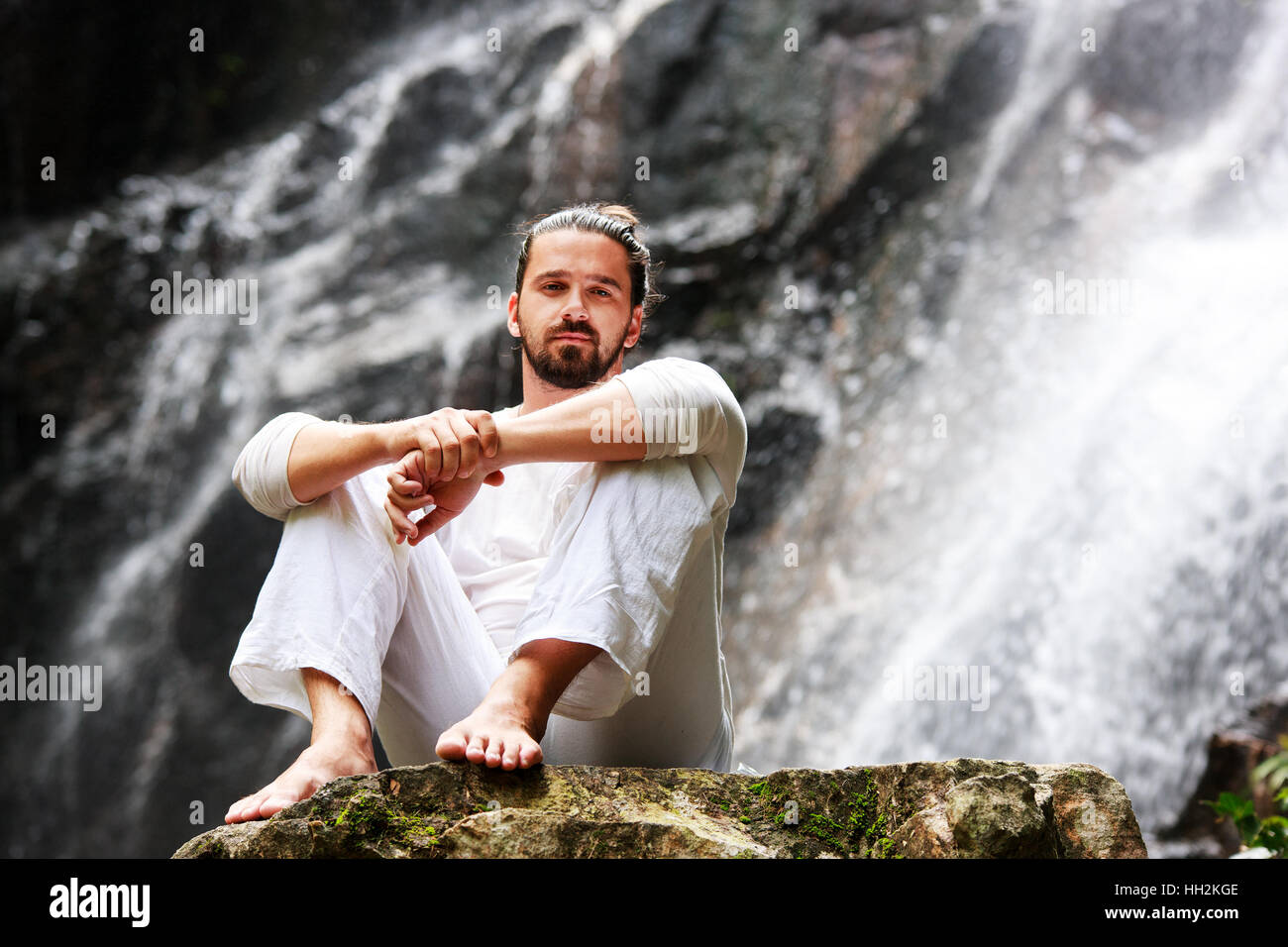 Mann sitzt in Meditation Yoga auf Felsen am Wasserfall im Regenwald Stockfoto