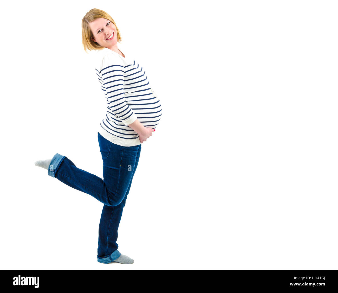 Blonde schwangere in Bluejeans und abgespeckte Pullover lächelnd und halten ihren Bauch mit einem Bein angehoben. Isoliert auf weißem Platz für Ihr Logo oder Stockfoto