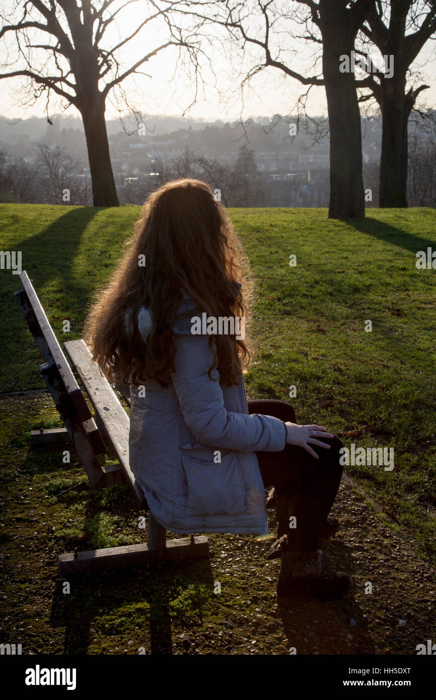 Rückansicht einer Frau allein sitzen auf einer Bank in einer ruhigen Lage mit einem hellen Himmel und Bäume. Stockfoto
