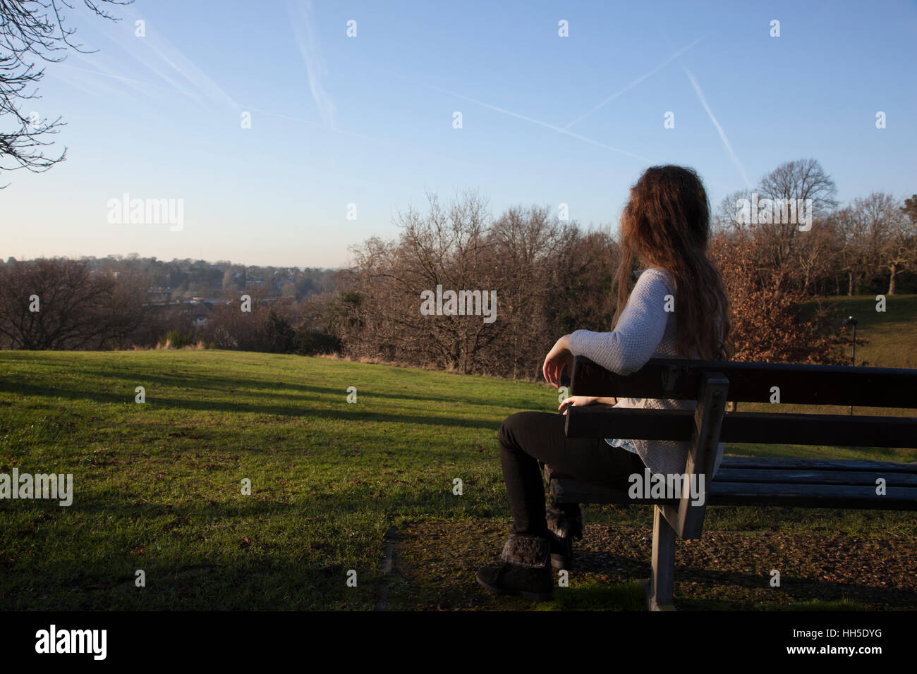 Nachdenkliche junge Frau sitzt allein auf einer Bank, Blick in die Ferne, in einer ruhigen Lage mit einem hellen Himmel und Bäume. Stockfoto