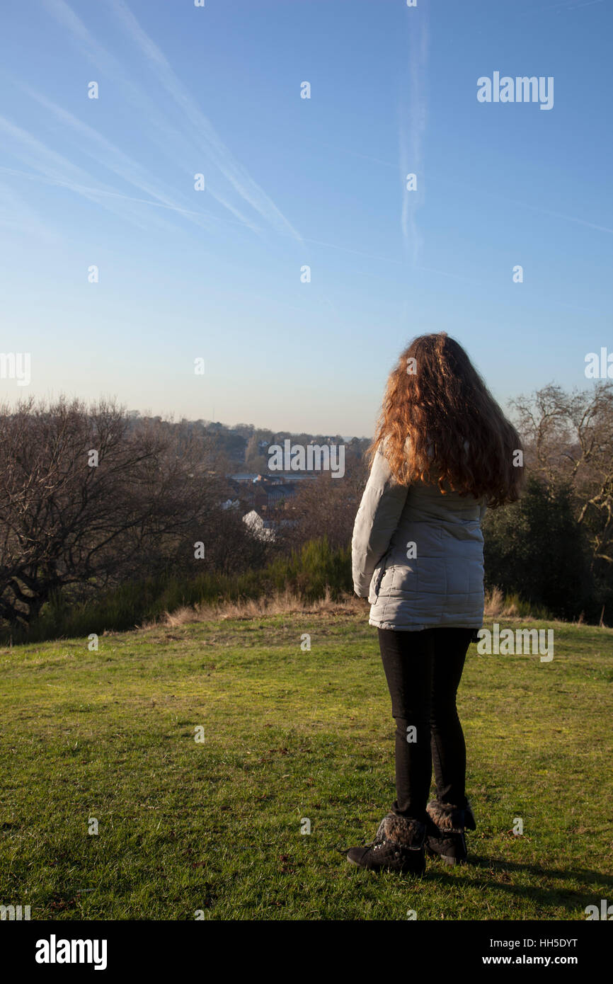 Nachdenkliche junge Frau allein stehend auf einem Hügel, der Blick in die Ferne, in einer ruhigen Lage mit einem hellen Himmel und Bäume. Stockfoto